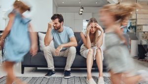 Deux parents assis sur un canapé ont l’air épuisés pendant que leurs enfants courent autour d’eux dans le salon. Scène réaliste de fatigue parentale et de perte de patience.